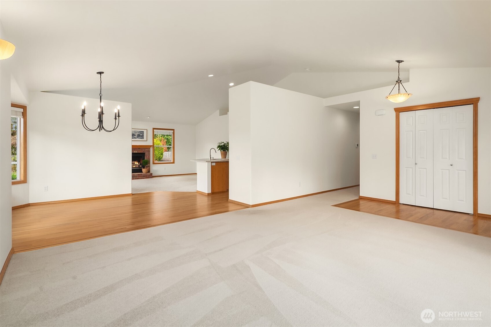 Empty living and dining area with cream carpet, hardwood floors, and a brick fireplace.