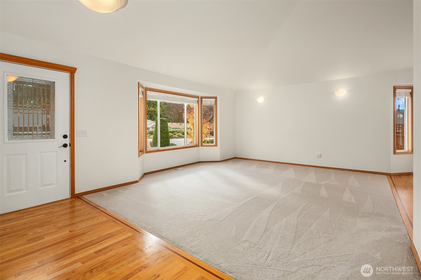 Unfurnished living room with a large pale carpet, hardwood flooring, and wood-trimmed windows.