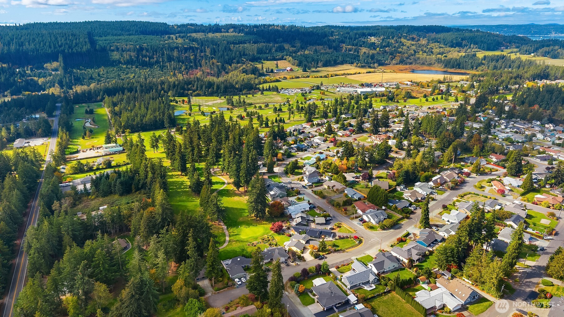 Aerial view of a neighborhood beside a wooded green space with winding paths and a distant pond.