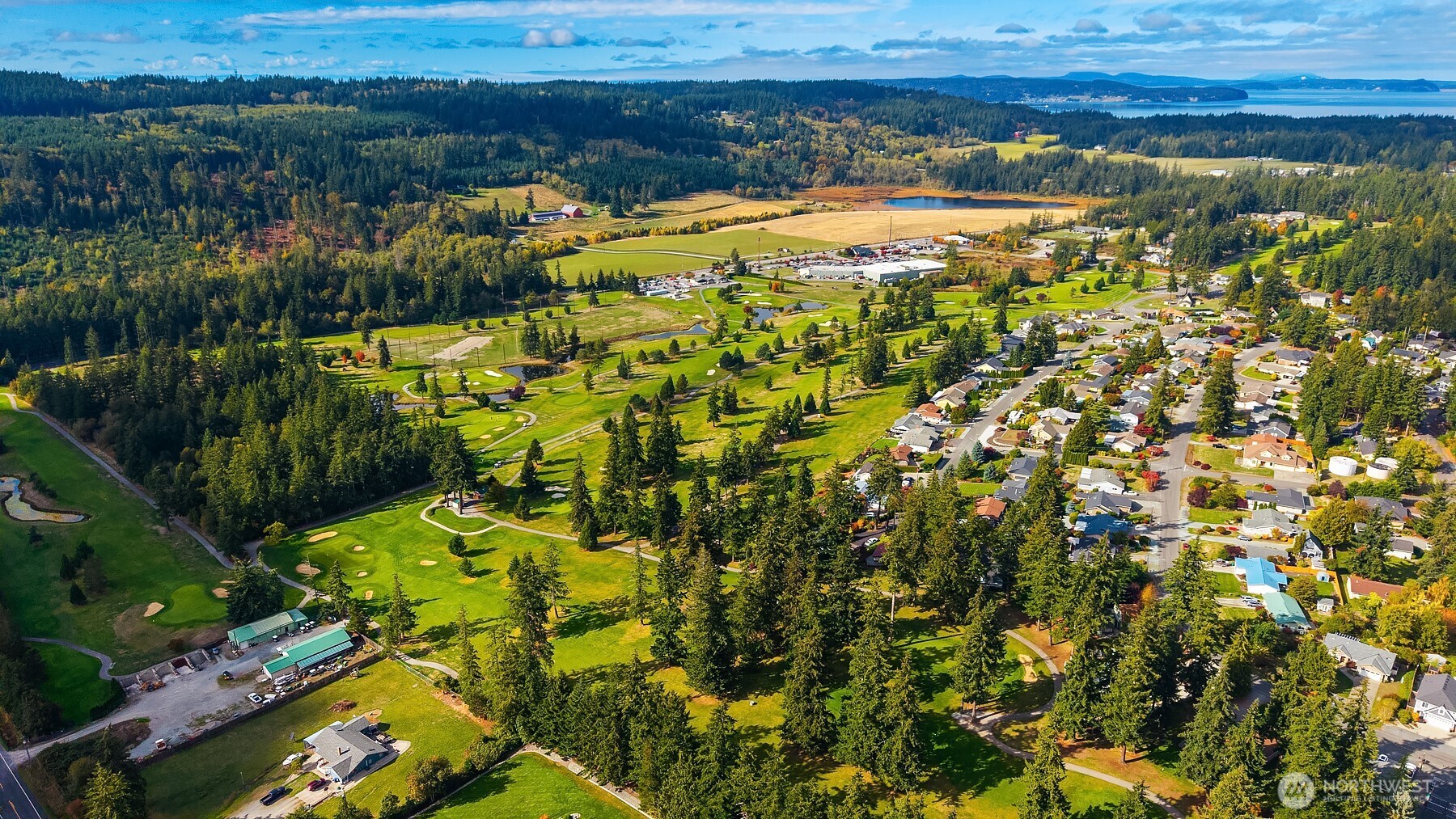 Aerial view of a golf course and nearby homes, with wooded hills and water in the distance.