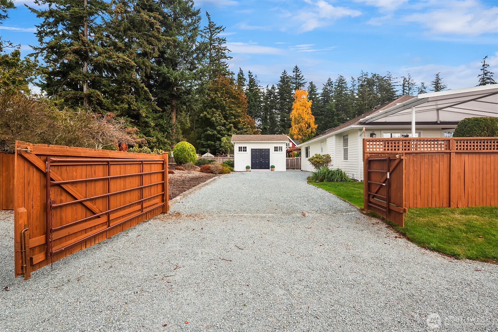 Gravel driveway through open wooden gates leading to a white house and detached outbuilding.