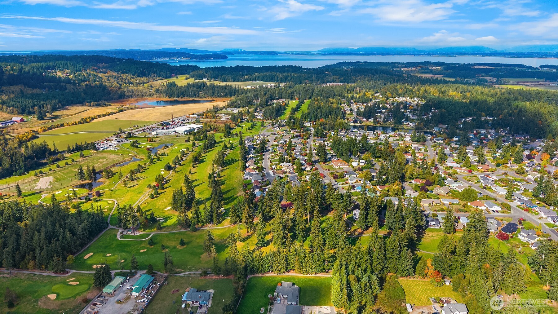 Aerial view of a golf course and neighborhood, with forest and water in the distance.