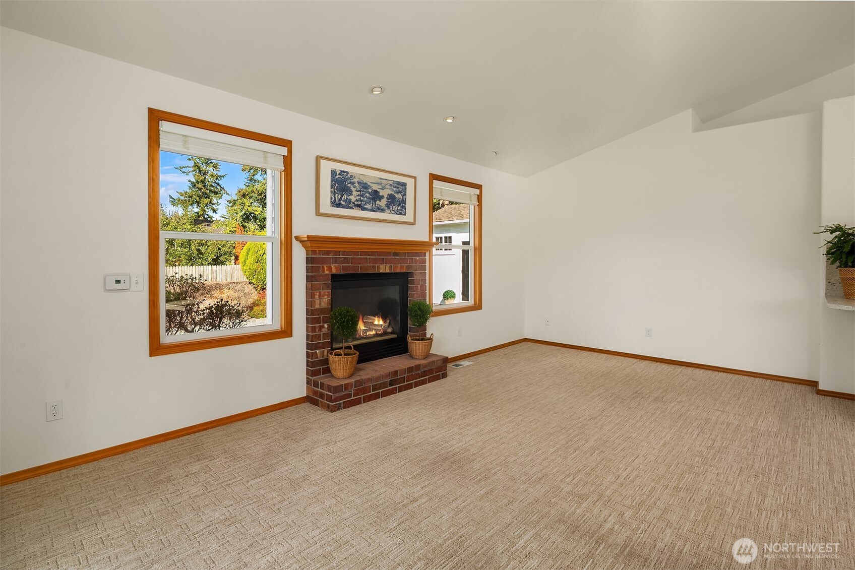 Empty living room with a brick fireplace, wood mantel, and two large windows.
