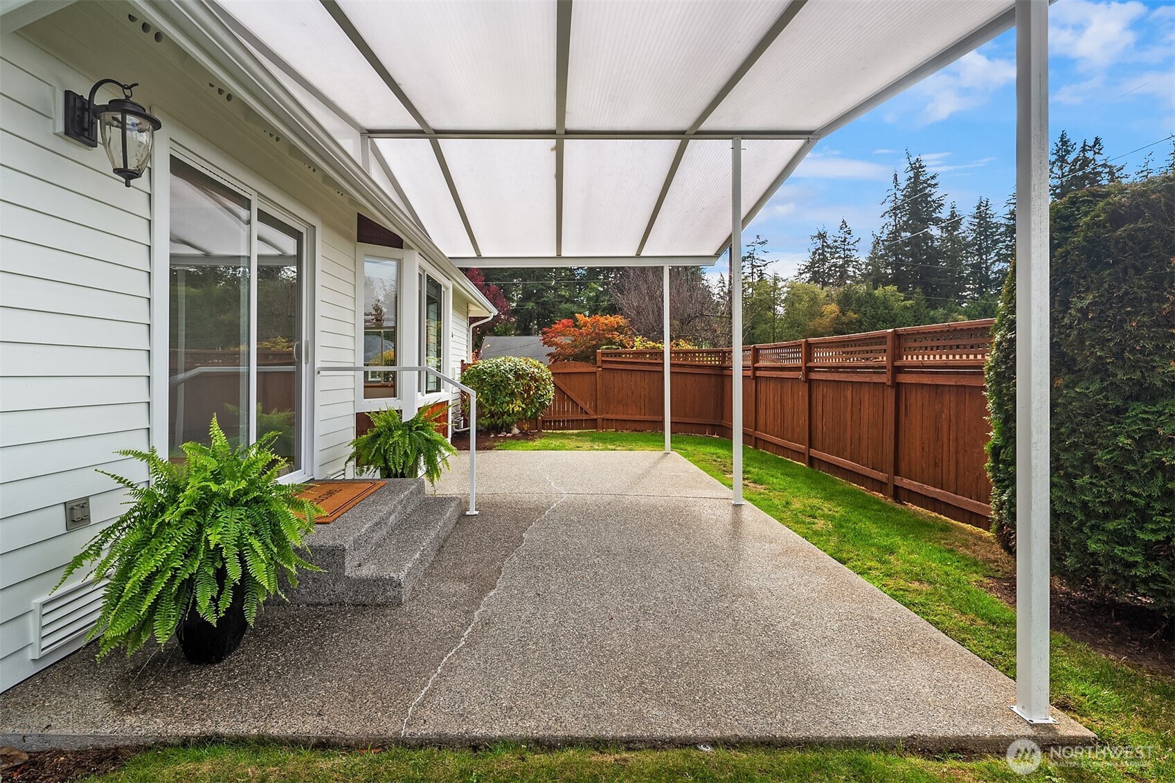 Covered concrete patio beside a light-colored house, with potted ferns and a fenced yard.