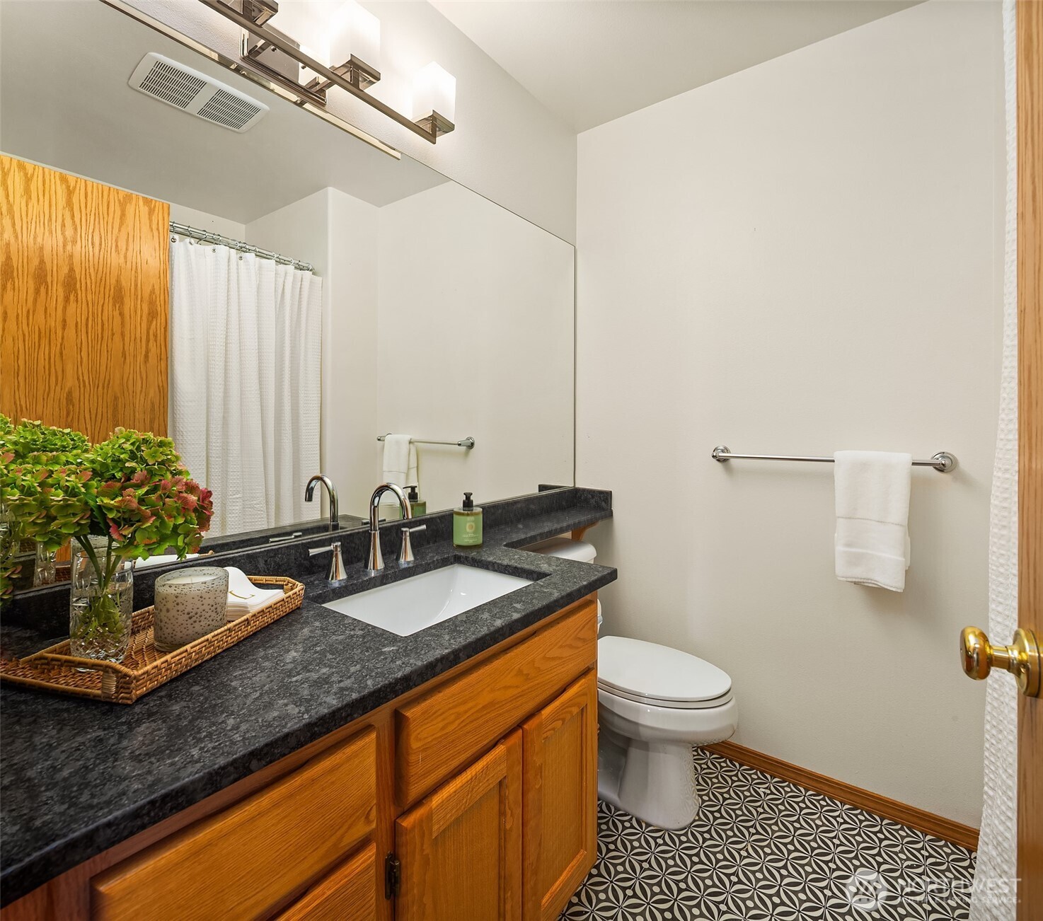 Bathroom with a wood vanity, dark stone counter, large mirror, and patterned tile floor