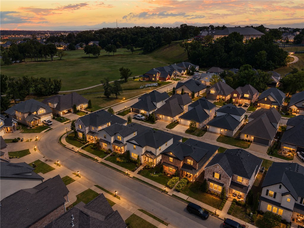 Aerial view of a neighborhood at dusk, with illuminated homes, curving streets, and a large grassy open space.