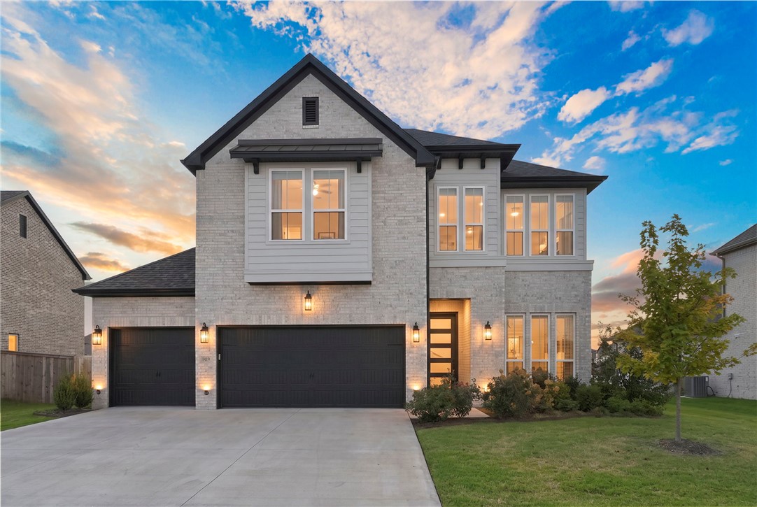 Two-story light-brick home with dark double garage doors and a wide driveway at golden hour.