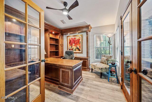 Home office with a built-in wood desk, floral artwork, and a cushioned chair beside tall windows.