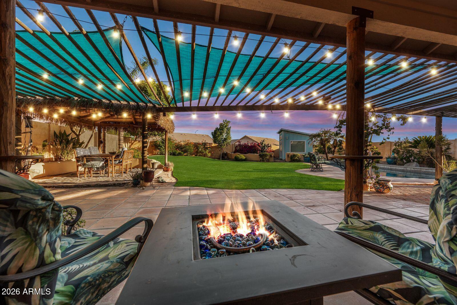 Backyard patio with a pergola, glowing fire pit, string lights, lawn, and swimming pool at dusk.