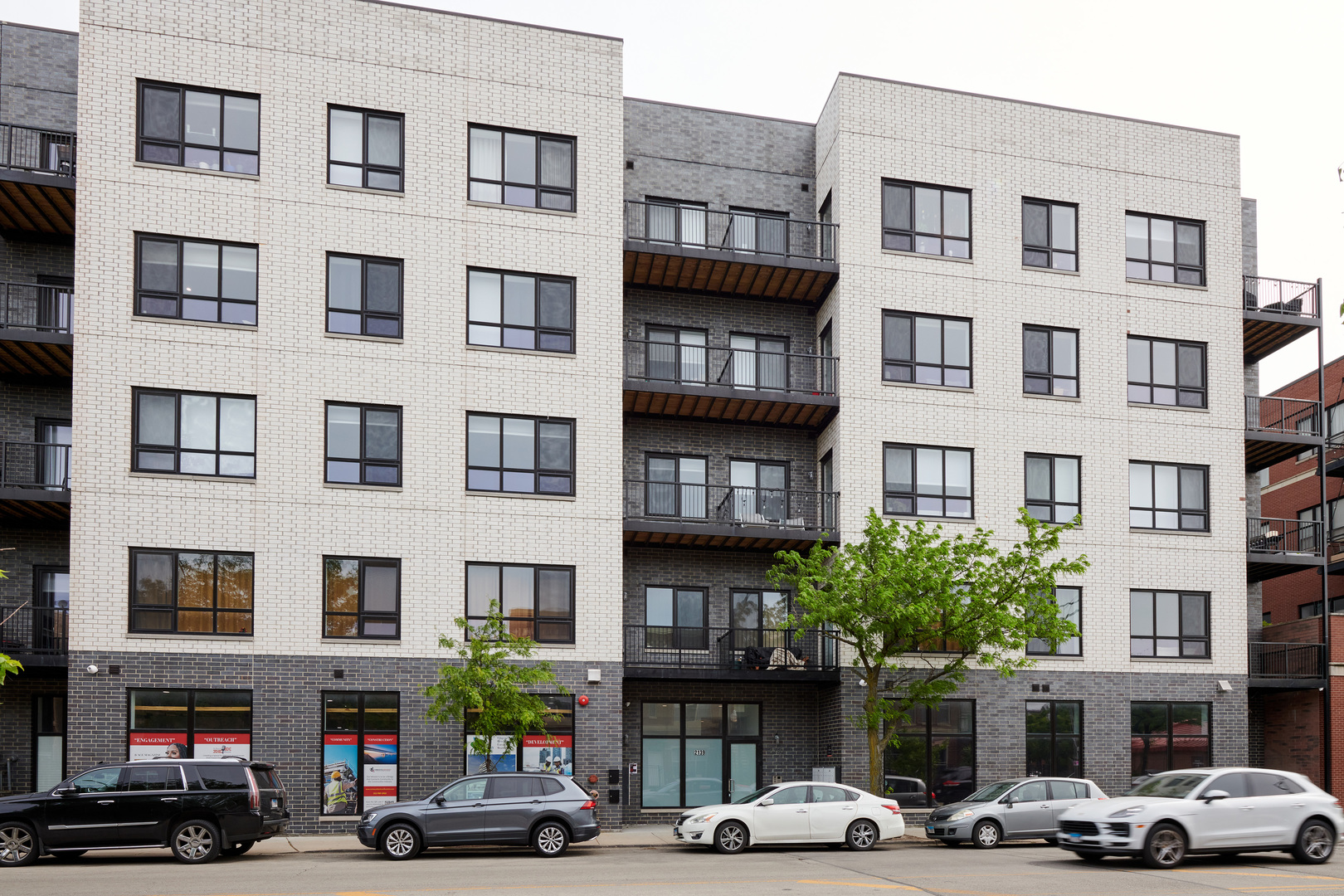 Modern white-brick apartment building with dark brick accents, balconies, and cars parked along the street.
