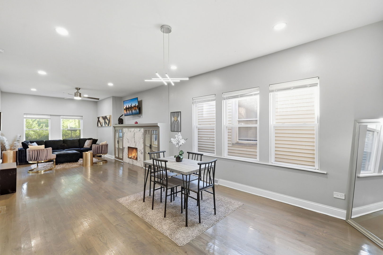 Open-plan living and dining room with a fireplace, black sofa, and dining table beside tall windows.
