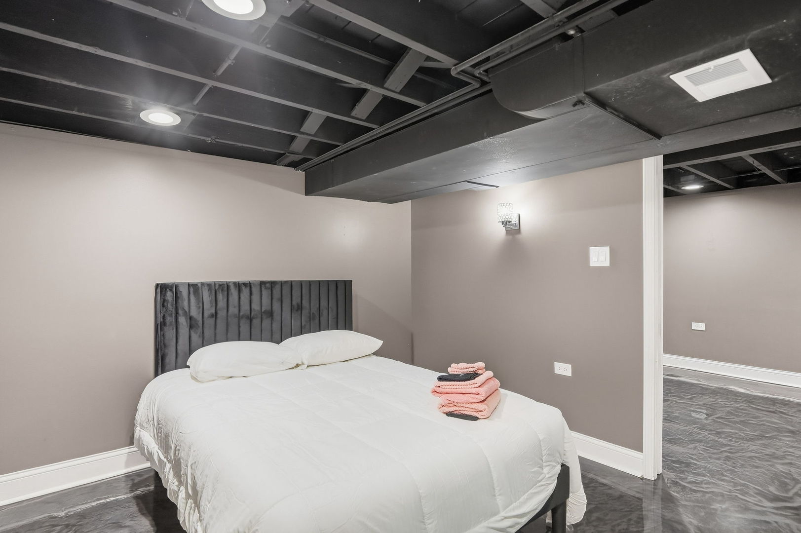Staged bedroom with a white bed, charcoal headboard, and exposed dark ceiling.