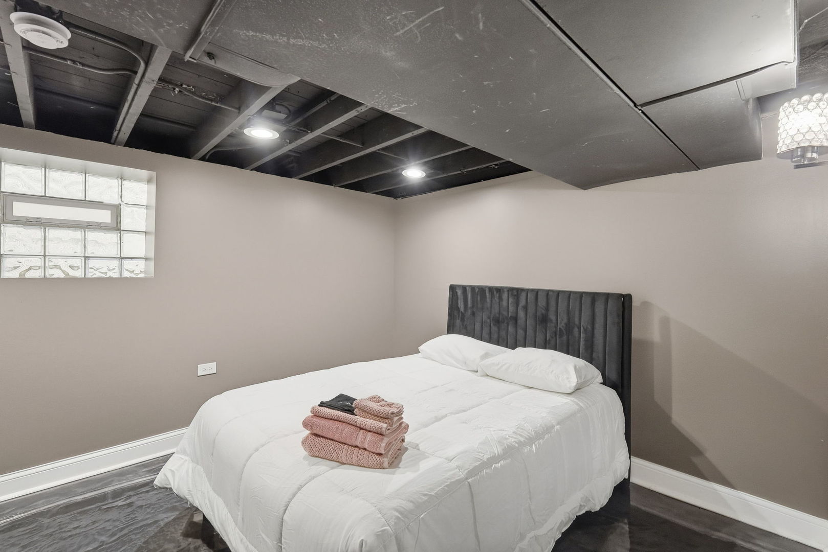 Basement bedroom with a white bed, dark upholstered headboard, glass-block window, and exposed ceiling.