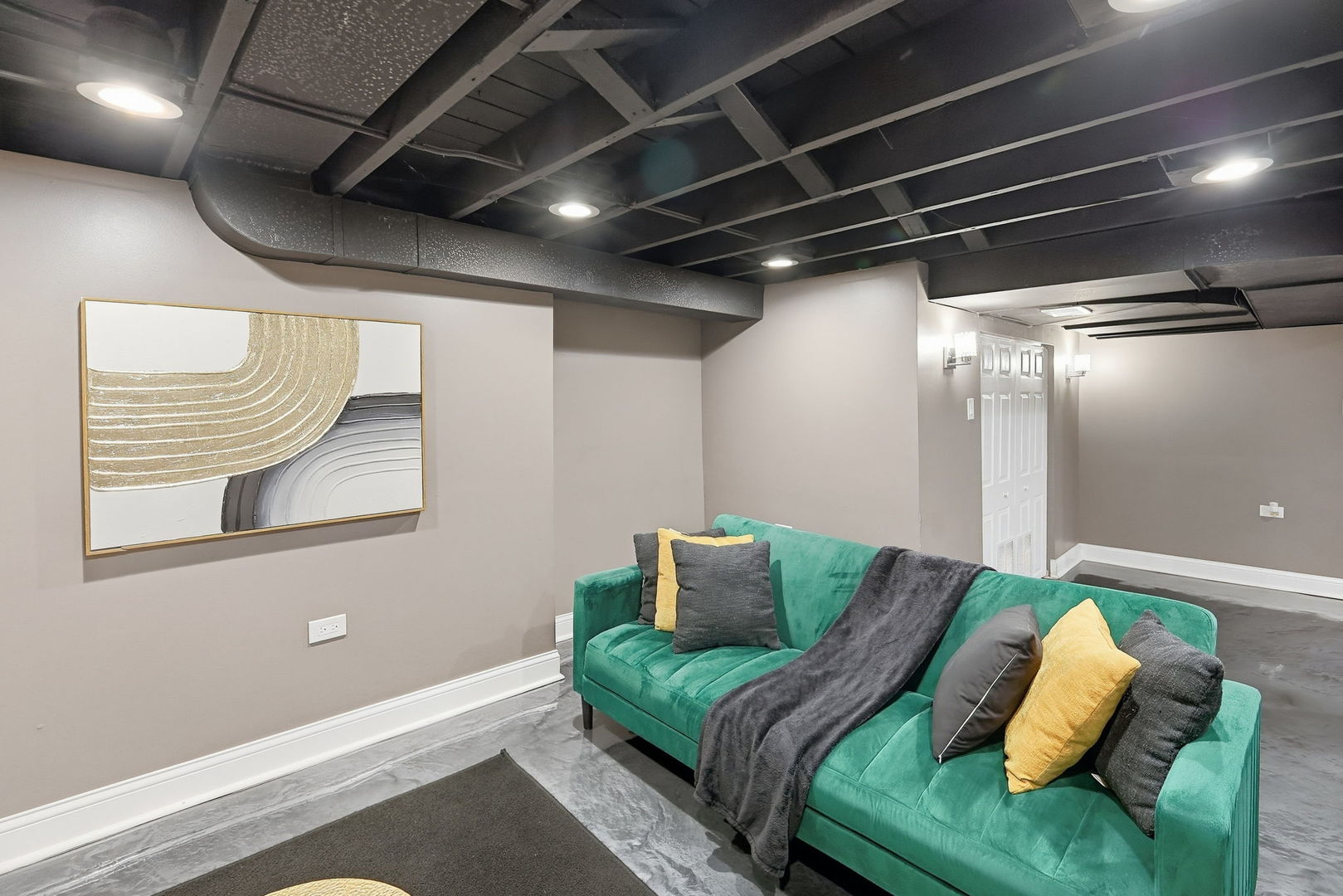 Finished basement lounge with a teal sofa, exposed ceiling joists, and polished concrete floor.