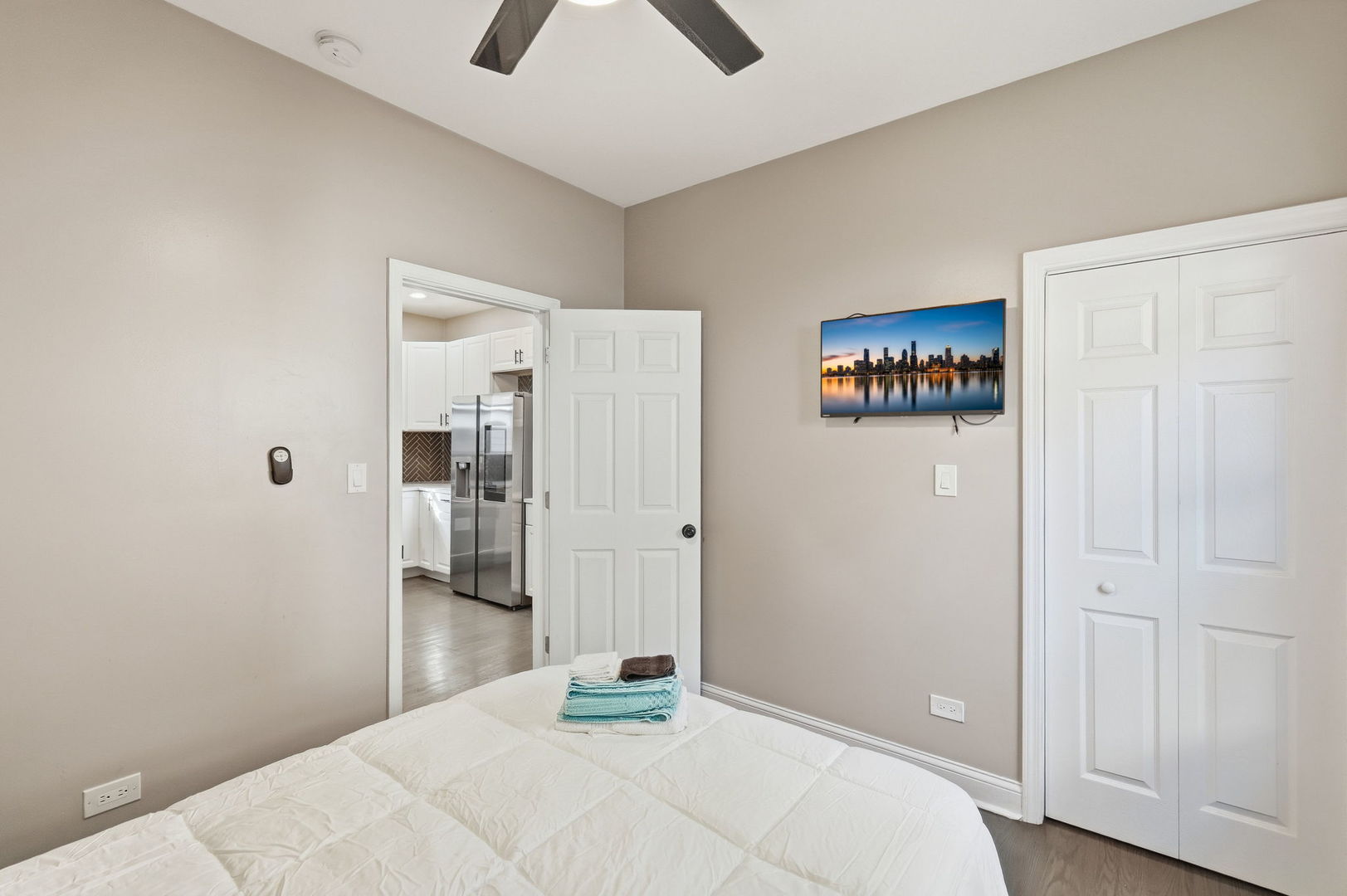 Bedroom with a white bed, ceiling fan, and open doorway to the kitchen.