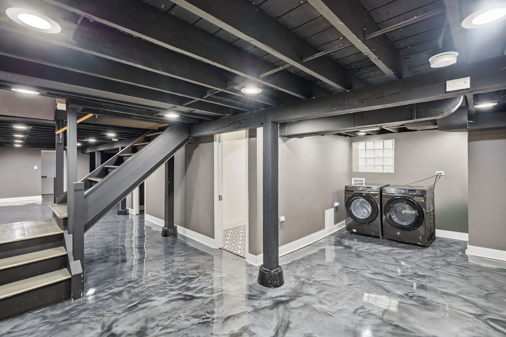 Basement laundry area with two front-loading machines beneath a glass-block window, beside a staircase.