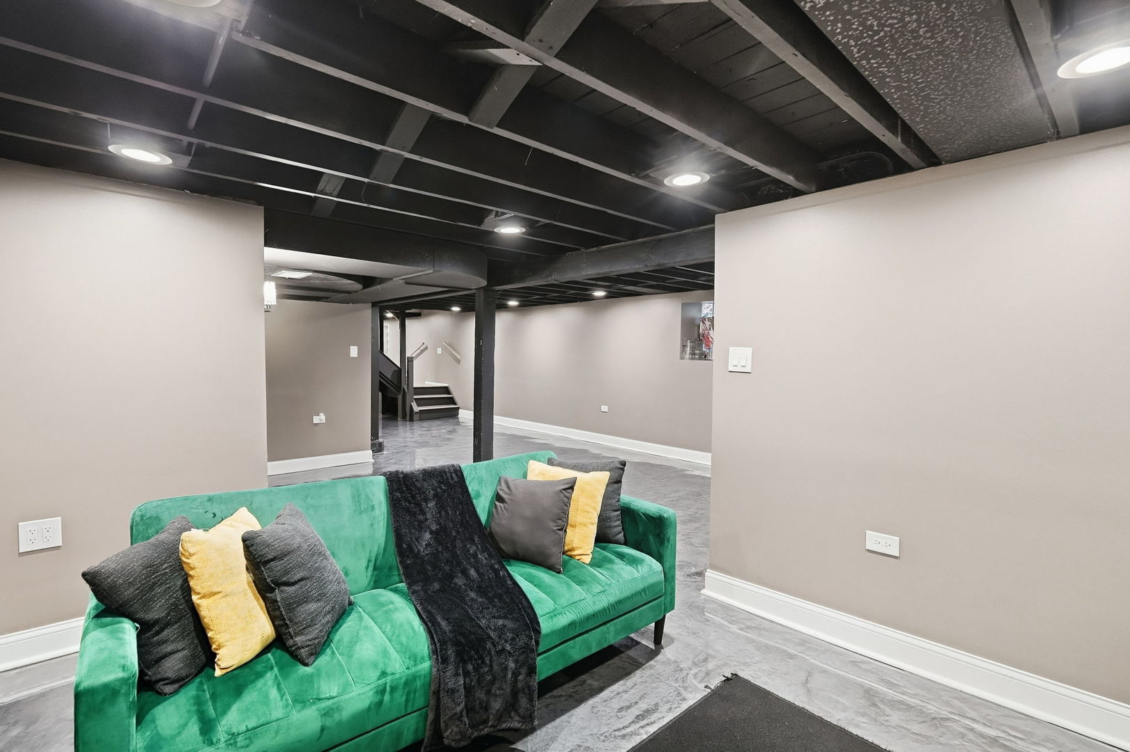 Finished basement living area with a green sofa, polished gray floor, and exposed black ceiling.