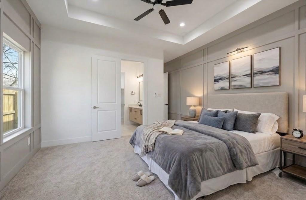 Staged bedroom with gray bedding, paneled walls, a tray ceiling, and an open doorway to the bathroom.