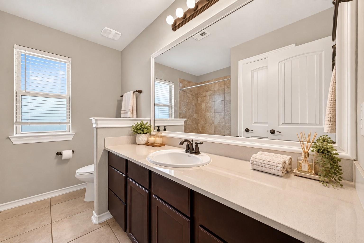 Bathroom with a dark wood vanity, long white counter, and tiled shower reflected in a large mirror.