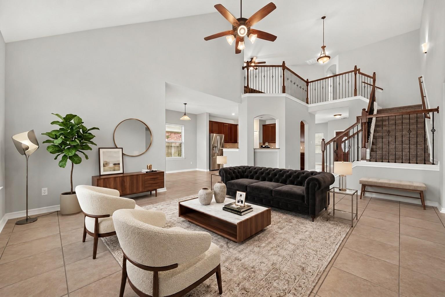 Two-story living room with a dark wood staircase, black sofa, cream chairs, and open kitchen.