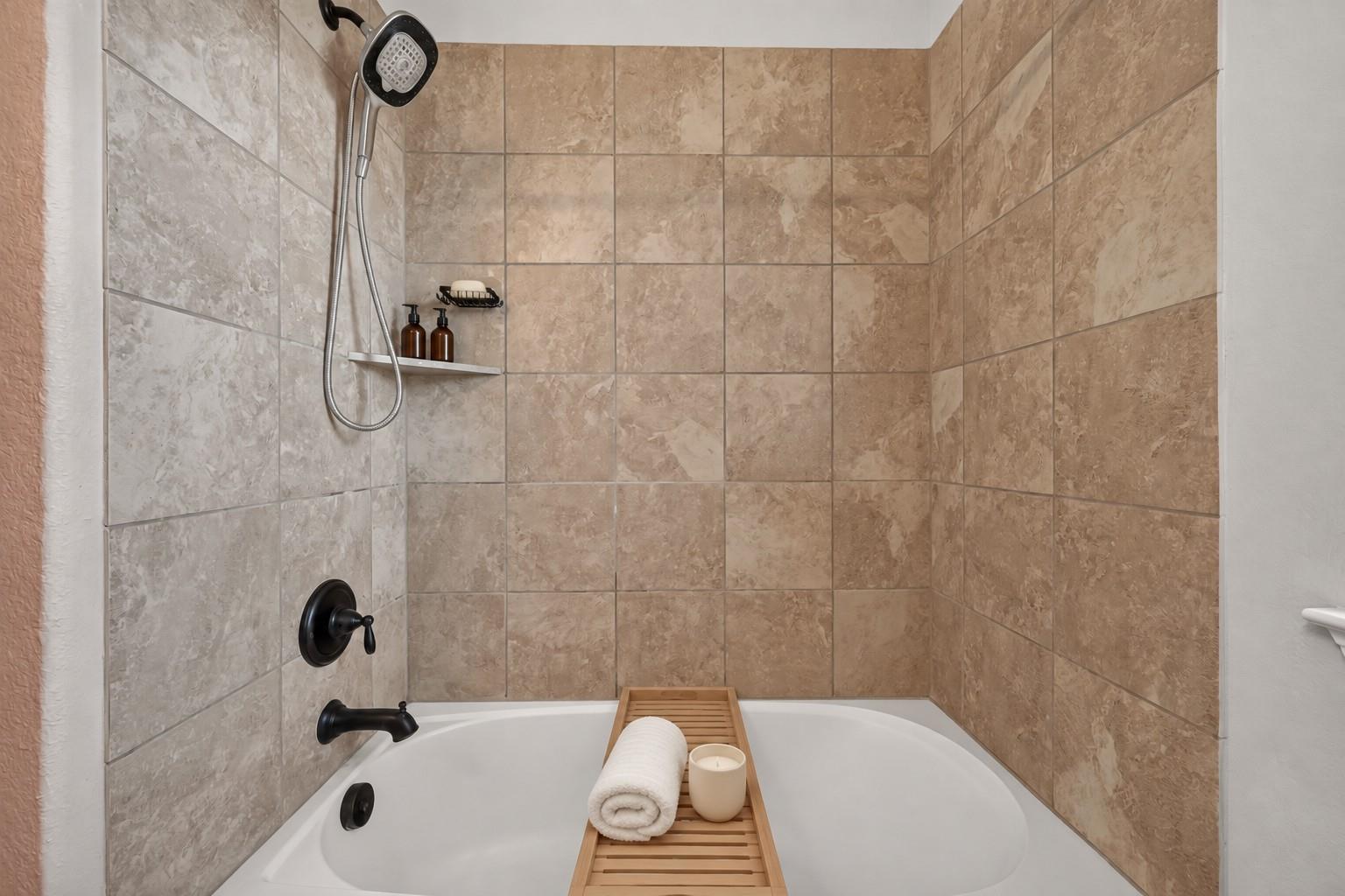 Bathroom bathtub and shower with beige tiled walls, dark fixtures, and a wooden tray with a towel and candle.