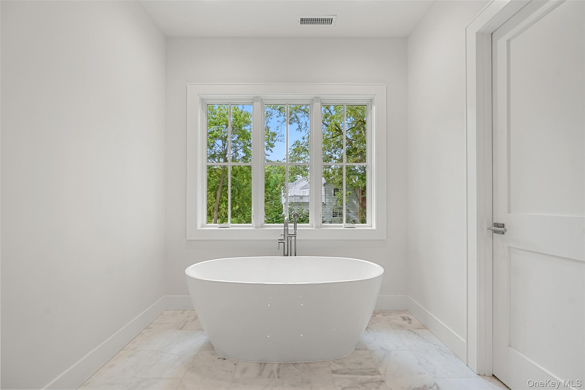 Bathroom with a freestanding soaking tub beneath a large window overlooking leafy trees.