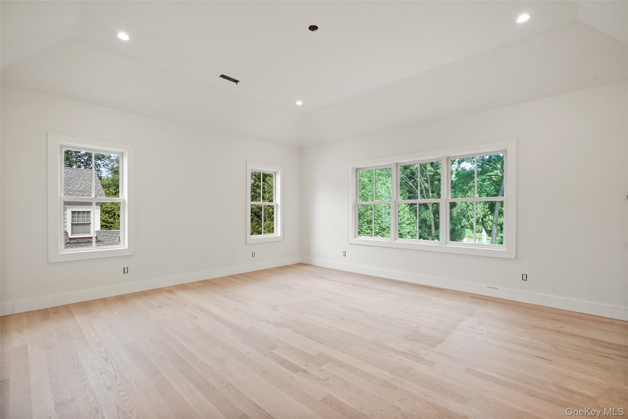 Bright empty room with pale hardwood floors and large windows overlooking trees.