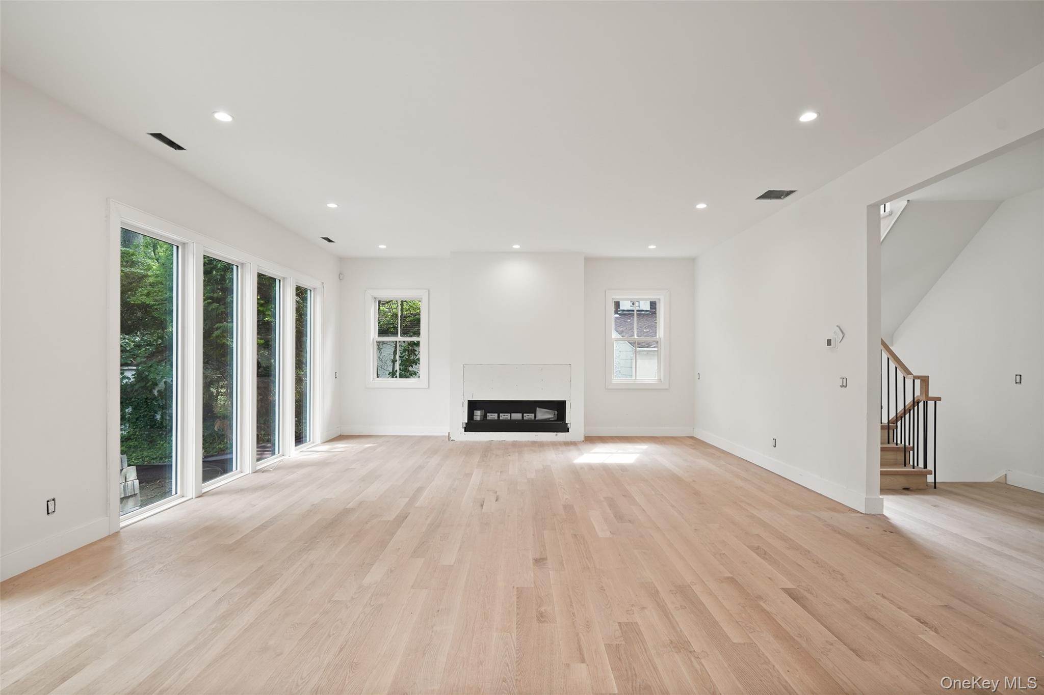 Vacant living room with pale hardwood floors, a central fireplace, tall windows, and an open staircase.