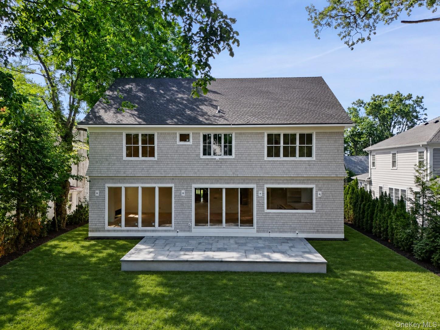 Rear of a gray-shingle house with a broad stone patio and neatly landscaped lawn.