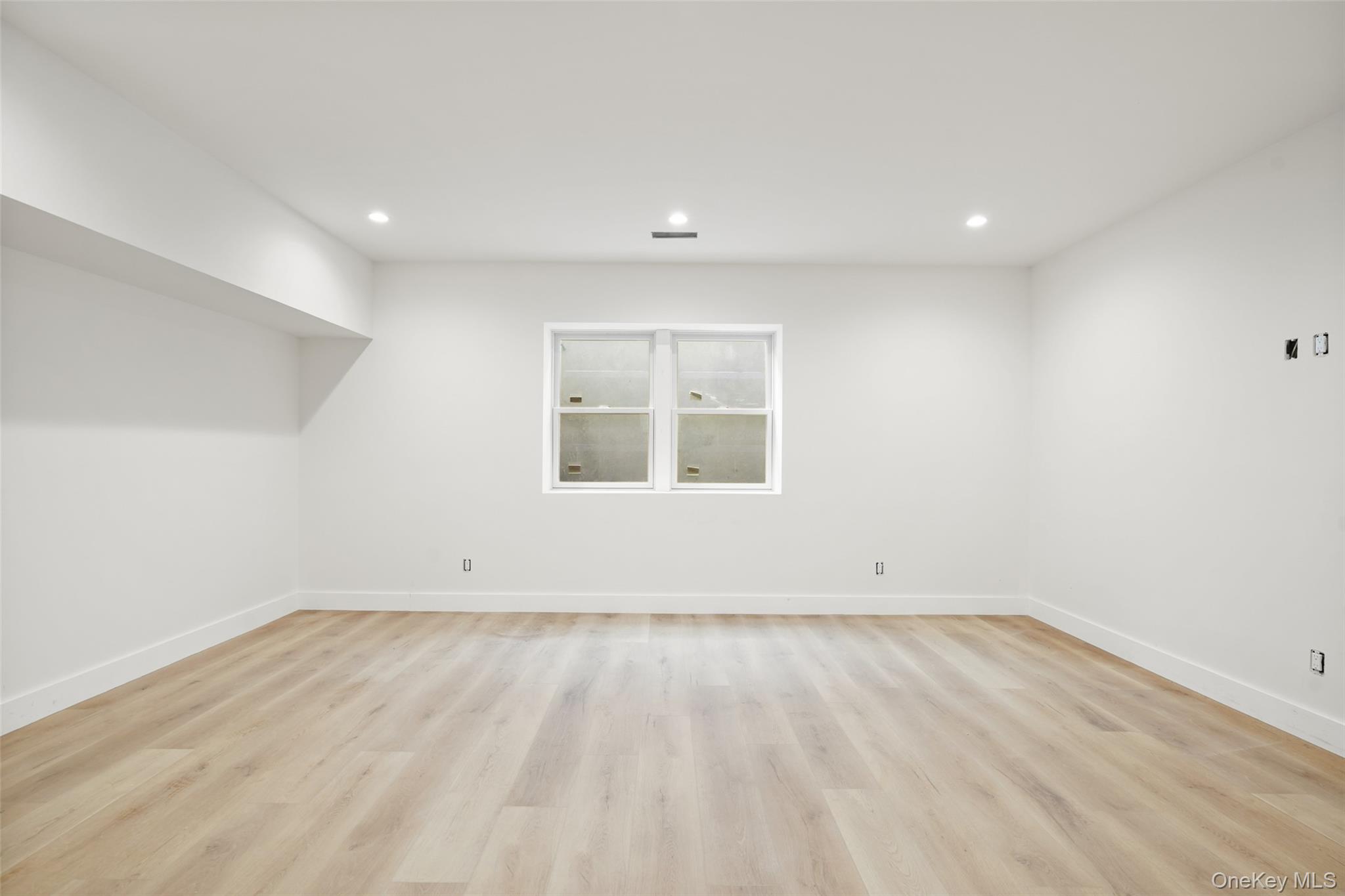 Empty room with pale wood flooring, recessed lights, and a centered window.