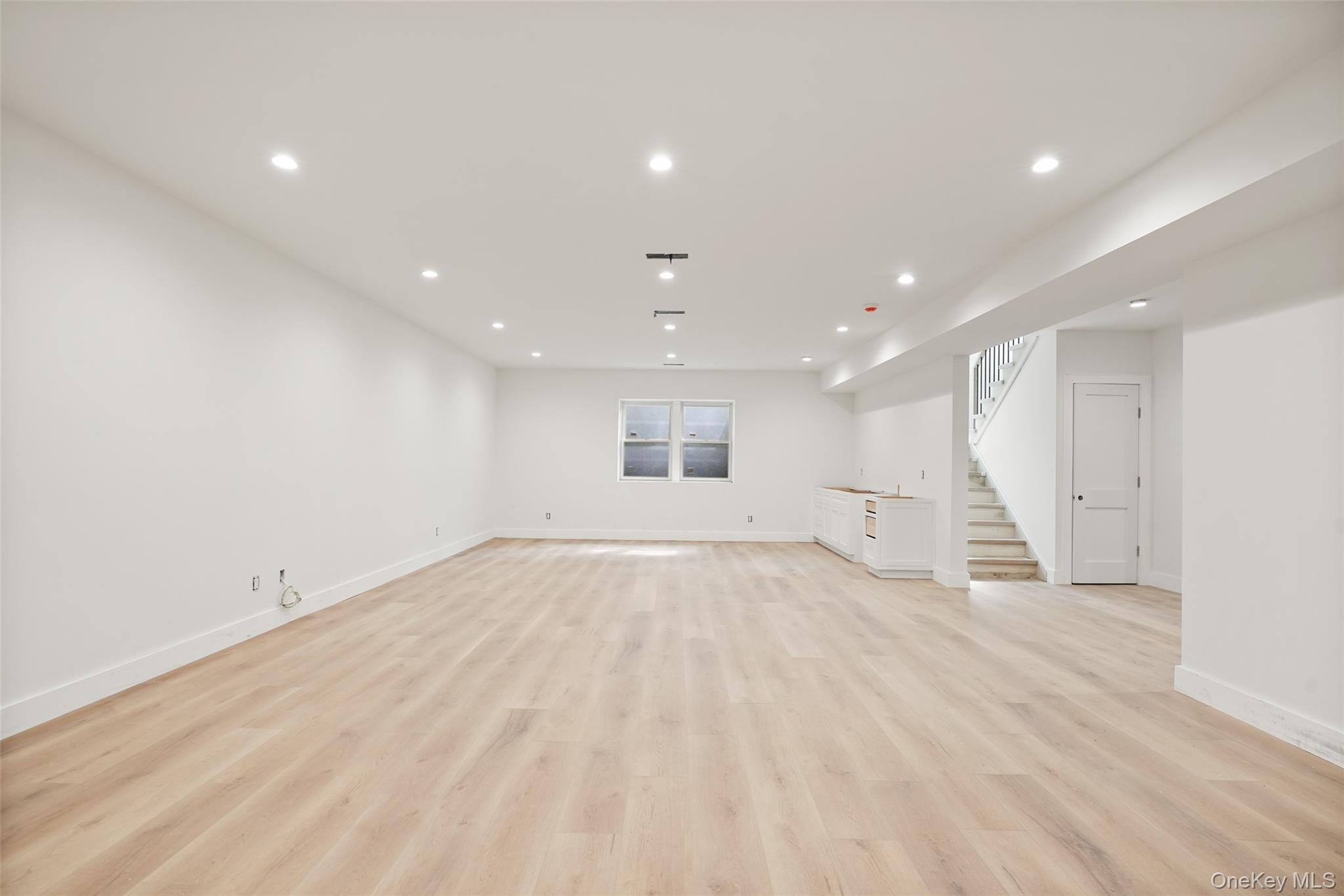 Vacant finished lower-level room with pale wood floors, recessed lights, built-in cabinetry, and stairs.