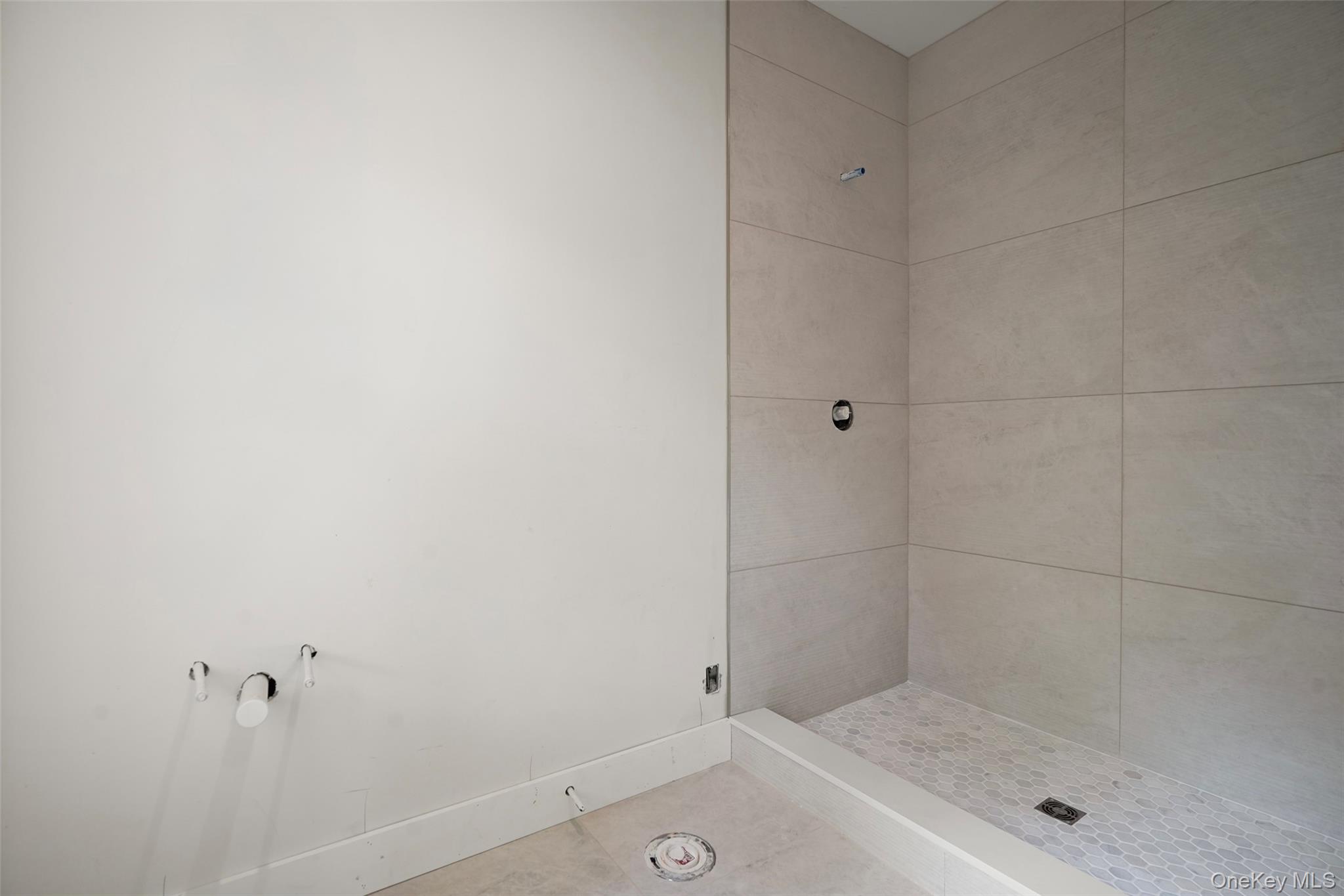 Unfinished bathroom shower with tiled walls, hexagonal floor, and exposed plumbing.