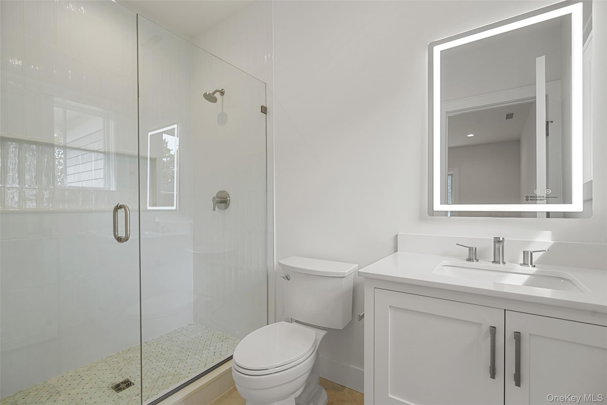 White bathroom with a glass walk-in shower, mosaic floor, and vanity with illuminated mirror.