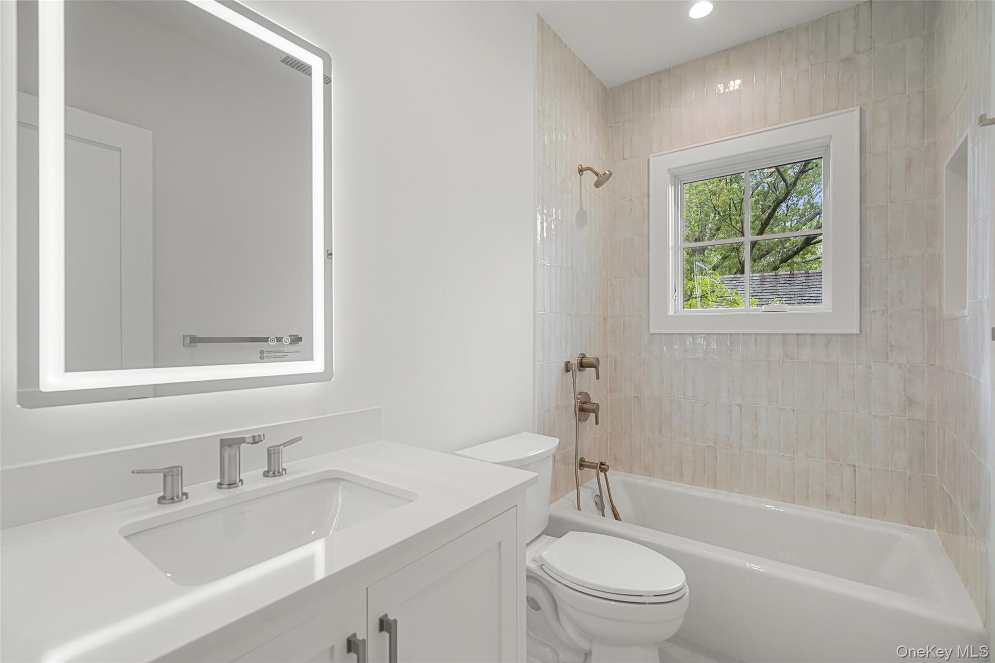 Bathroom with a white vanity, tiled tub surround, and window overlooking leafy trees.