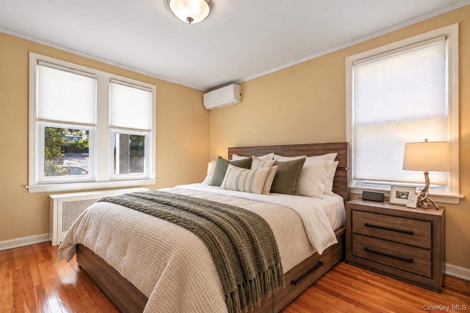 Staged bedroom with a wood bed, layered neutral bedding, hardwood floors, and shaded windows.