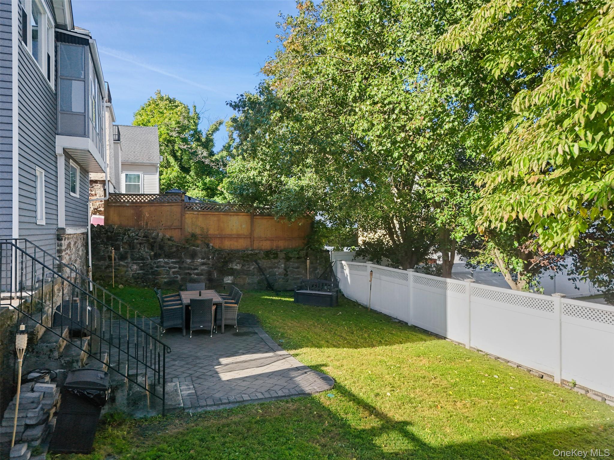 Backyard with a paved patio and dining set, green lawn, mature trees, and fenced boundaries.