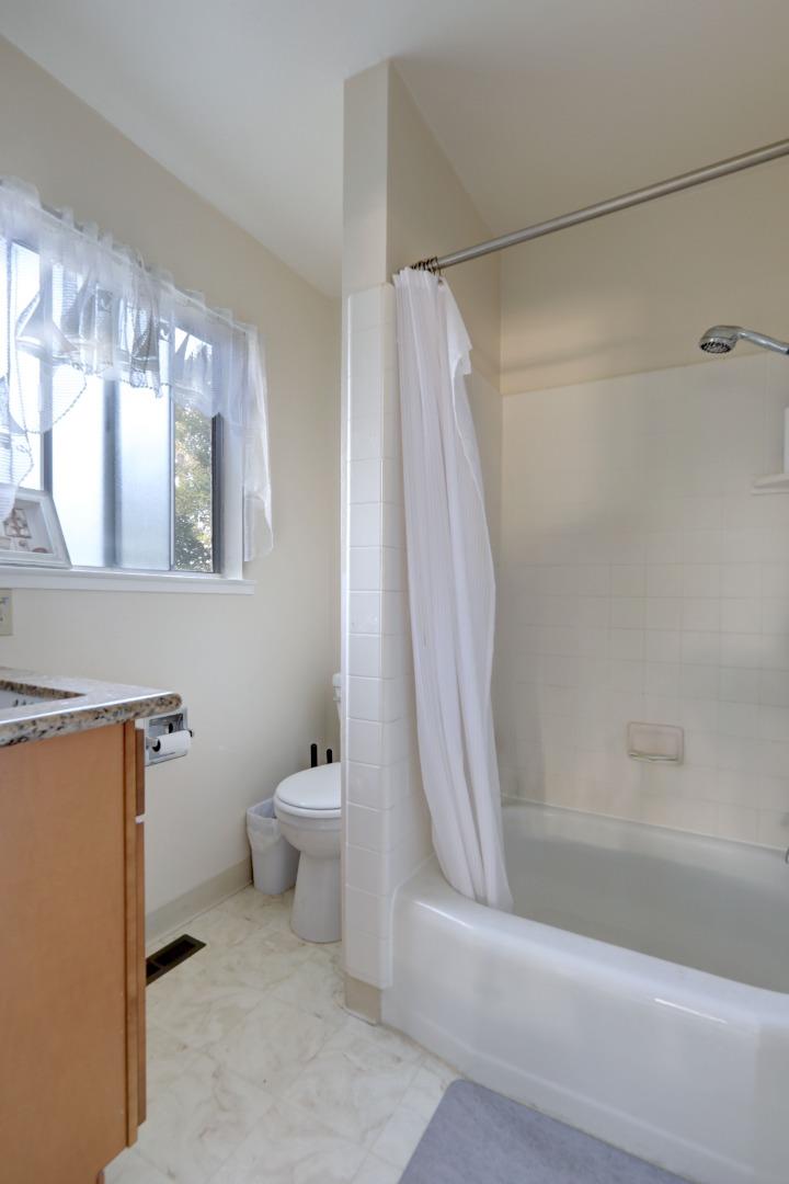 Bathroom with a curtained bathtub and shower, toilet, wood vanity, and lace-curtained window.