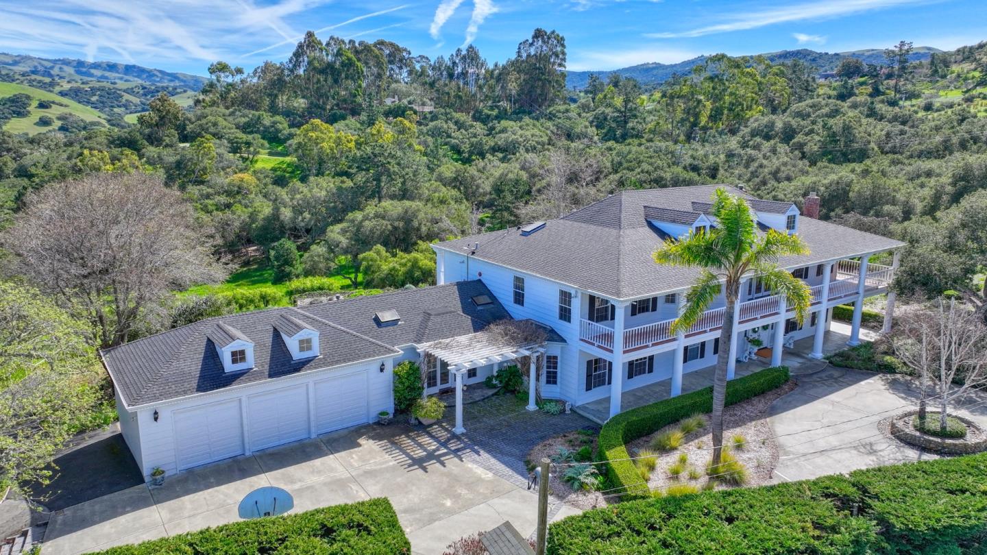 Aerial view of a white two-story home with a wraparound balcony, surrounded by wooded hills.