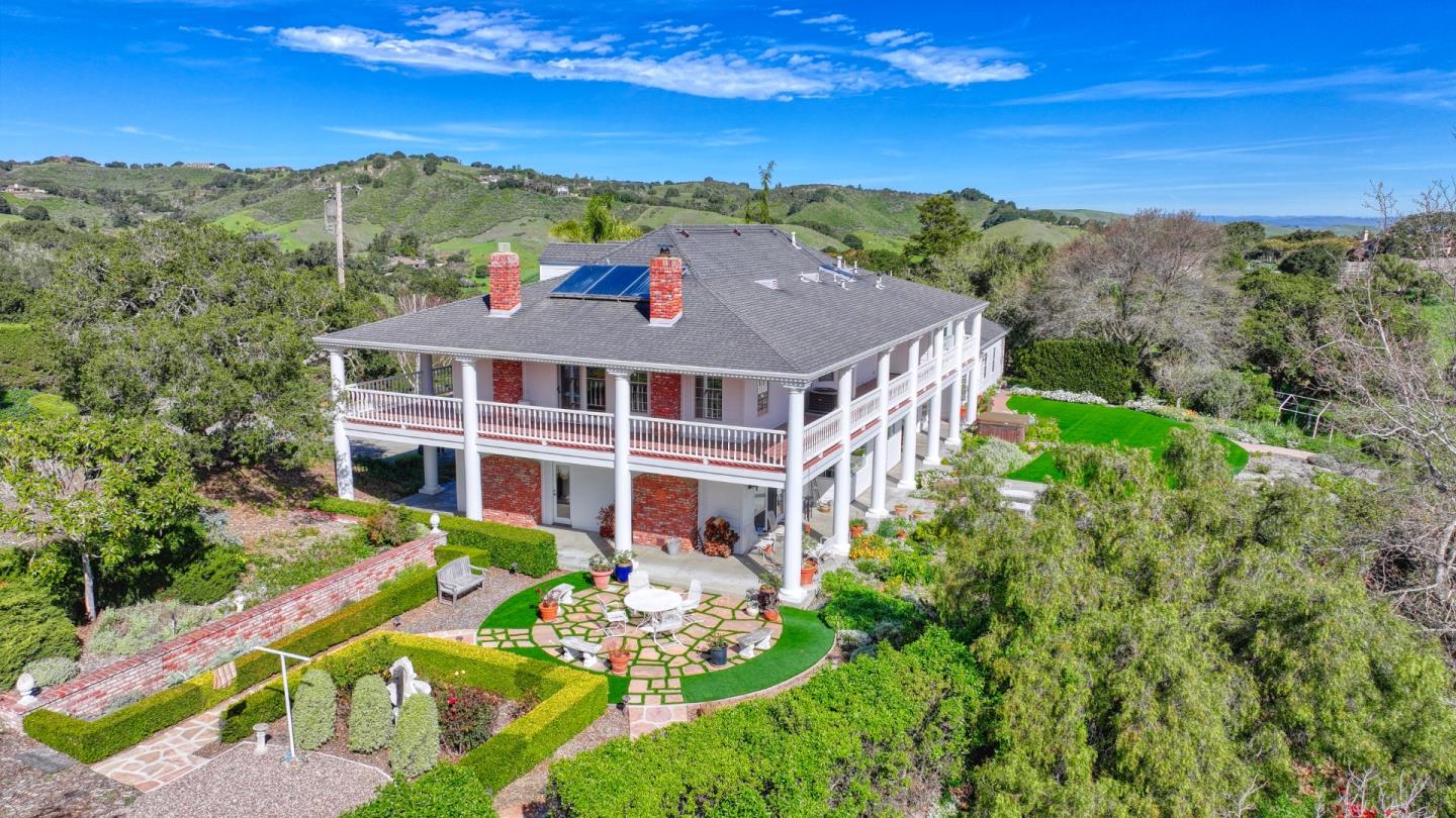 Aerial view of a two-story brick-and-white-column home with wraparound balconies, patio, and rolling green hills.
