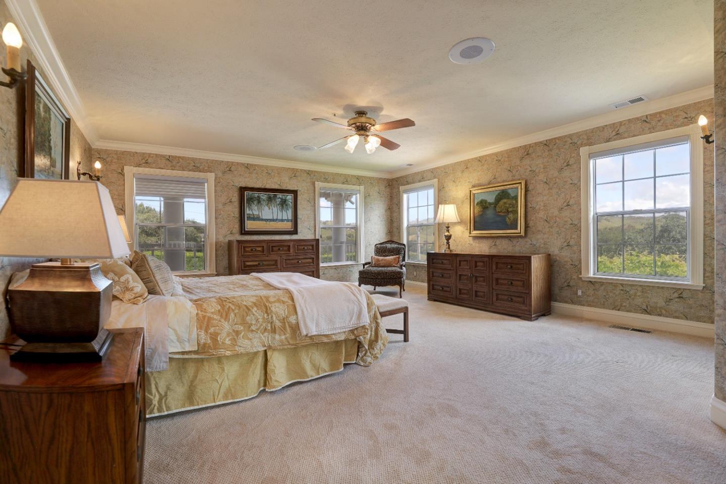 Furnished bedroom with patterned wallpaper, wooden dressers, a ceiling fan, and large windows overlooking greenery.
