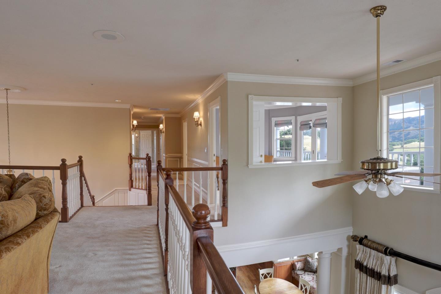 Carpeted upstairs landing overlooks a living room, with wood railings, a ceiling fan, and hills beyond the windows.