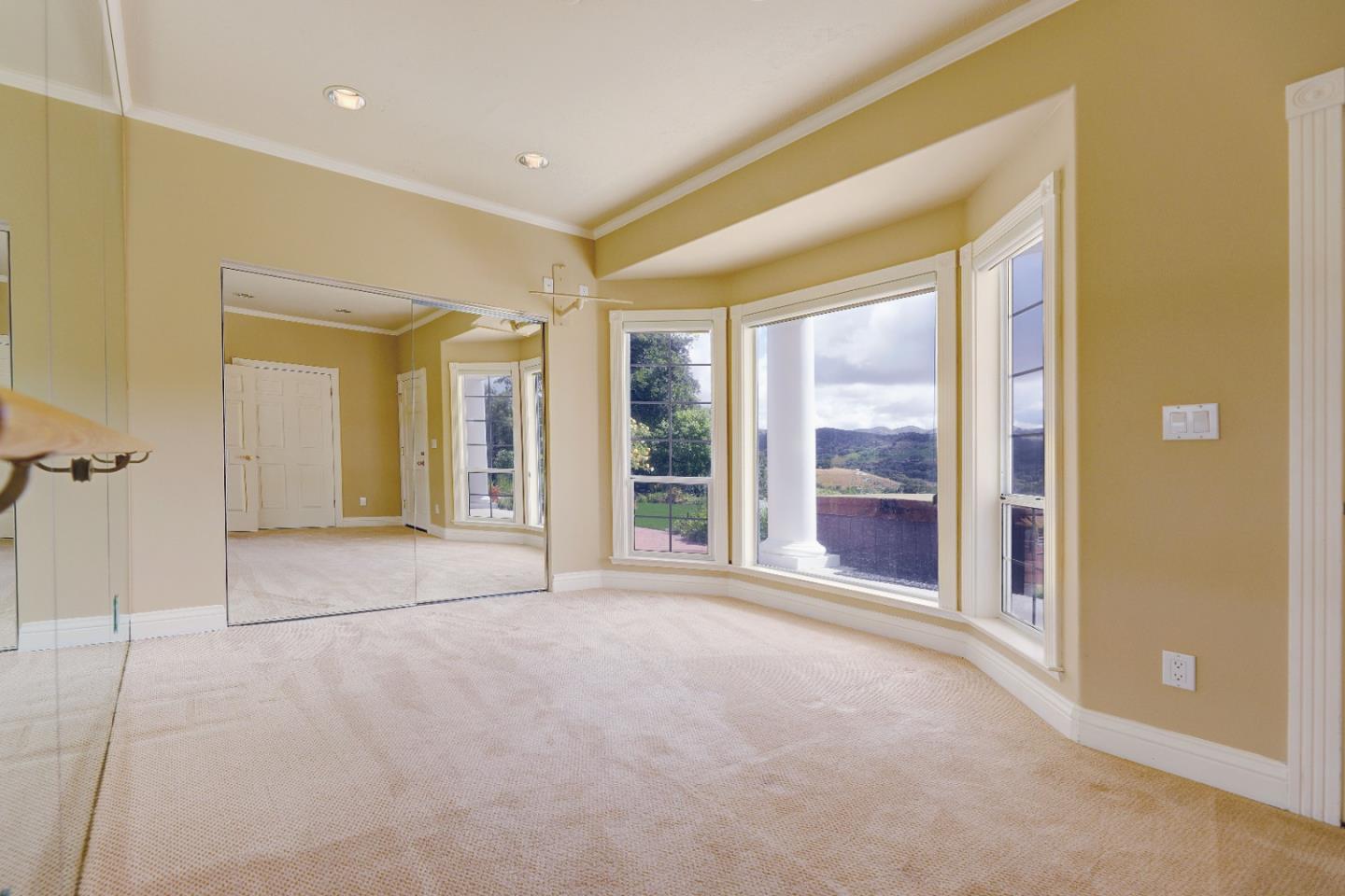 Empty carpeted bedroom with mirrored closet doors and tall windows overlooking hills.