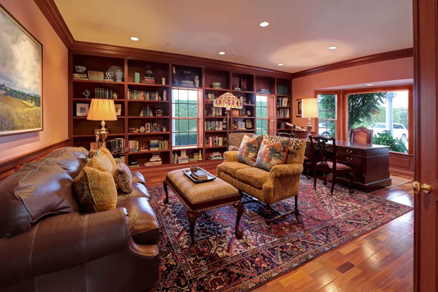 Furnished library with built-in bookshelves, leather sofa, patterned rug, and a desk beside large windows.
