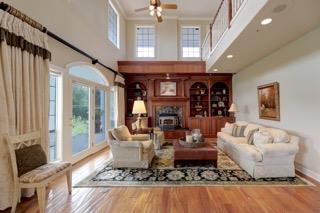 Two-story living room with a fireplace, built-in shelves, cream sofas, and a balcony overlooking tall windows.