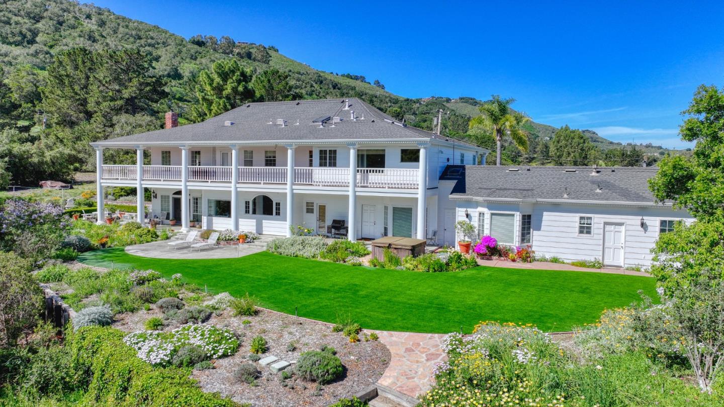 Rear exterior of a two-story white home with balcony, landscaped lawn, and mountain backdrop.
