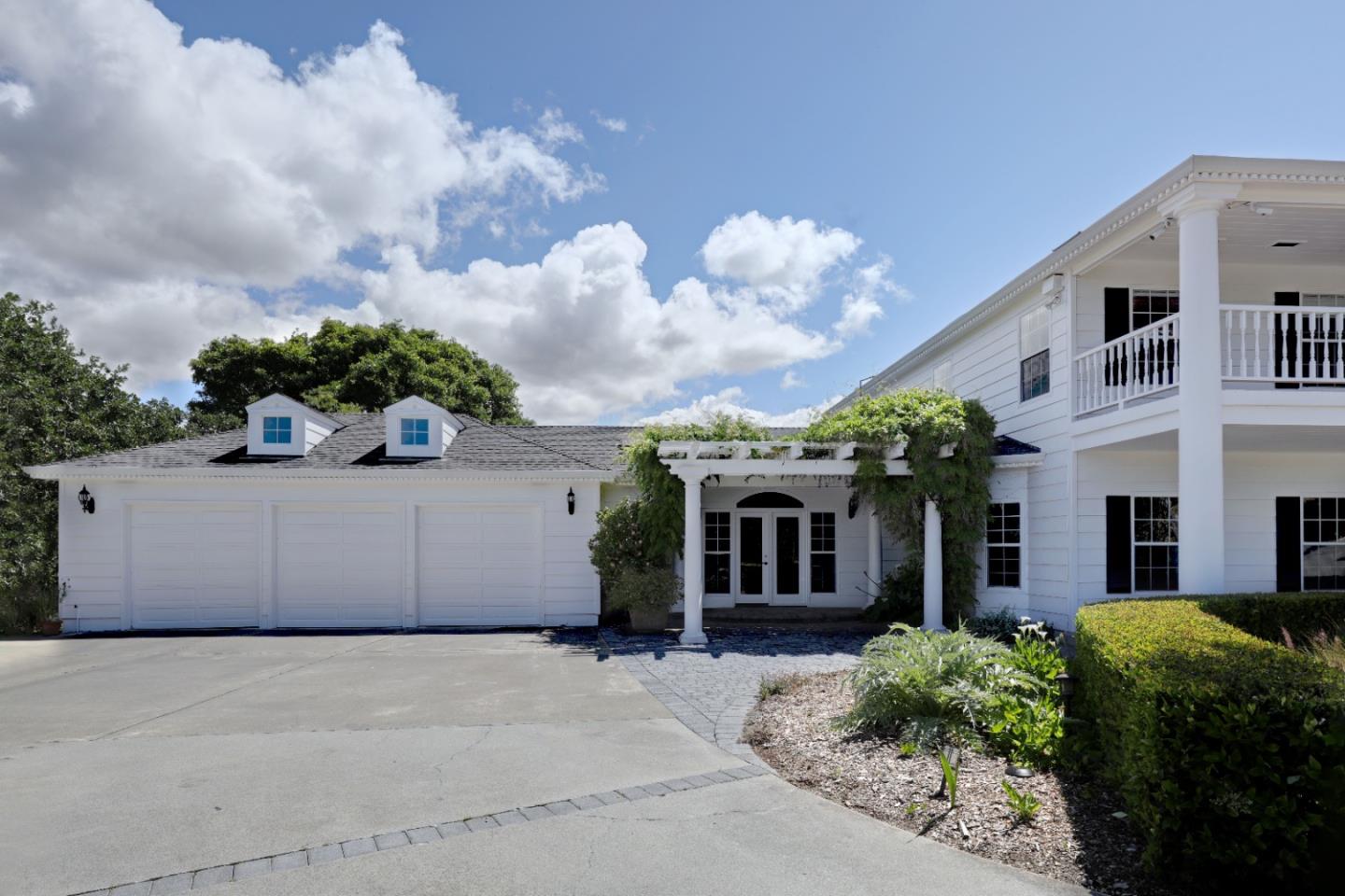 White house with a three-car garage, vine-covered entry pergola, upper balcony, and broad driveway.