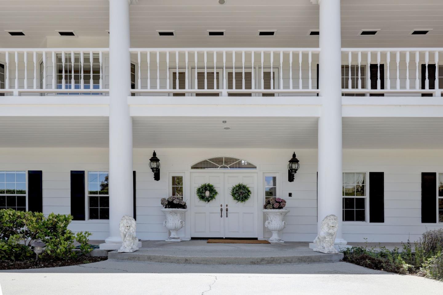 White two-story home entrance with tall columns, balcony, wreath-adorned doors, and stone lion statues.