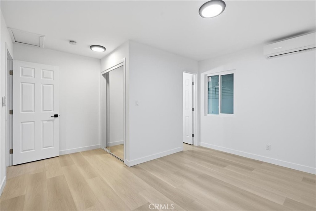 Empty room with light wood floors, mirrored closet doors, a window, and a wall-mounted air conditioner.