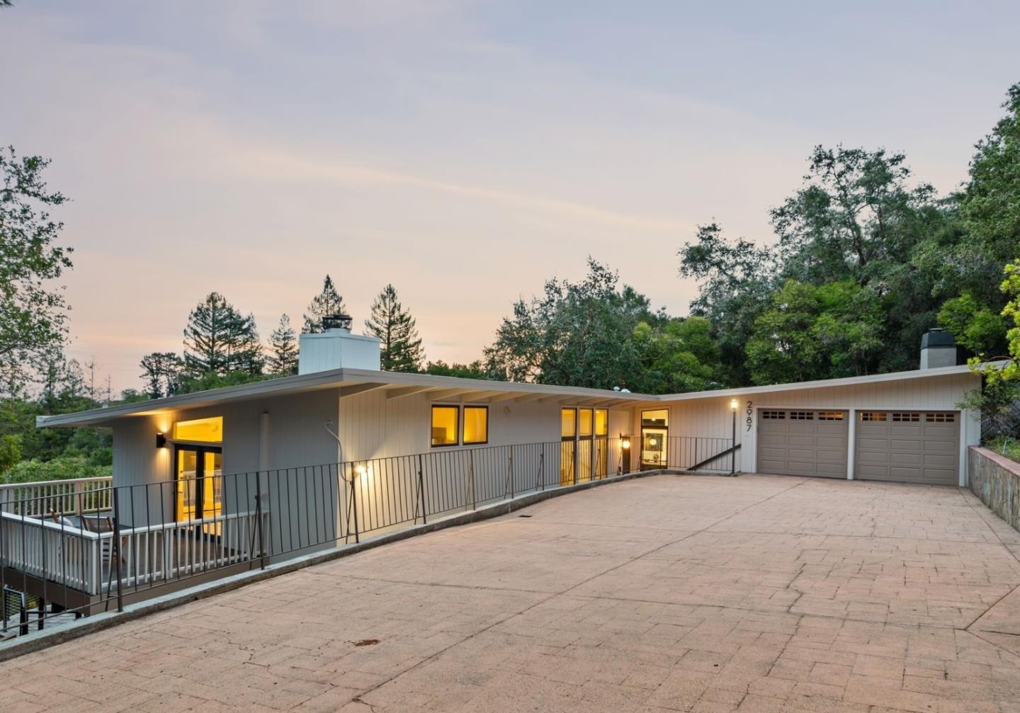 Contemporary home with a broad paved driveway, two-car garage, and wooded surroundings at dusk.