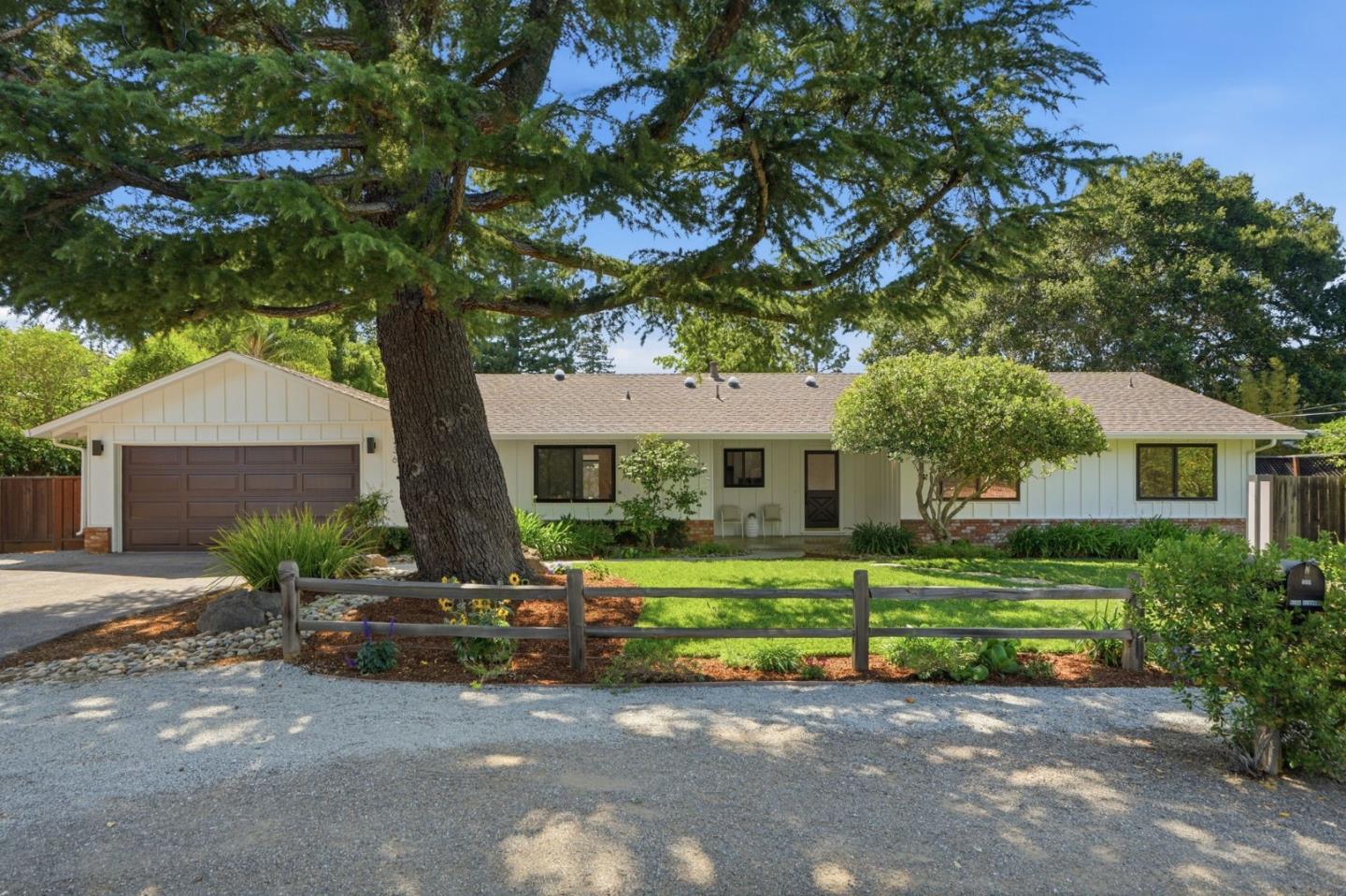 Single-story white house with a brown garage, mature shade tree, and low wooden fence.